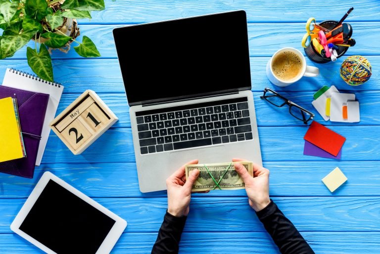 hands holding money by laptop on blue wooden table with stationery